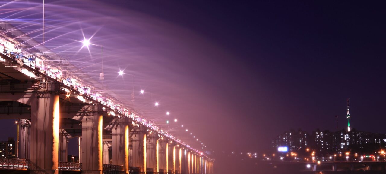 Night view of an illuminated bridge and city skyline, showcasing vibrant lights and urban architecture. 汉江野餐团, 盘浦汉江公园野餐垫, Some Sevit夜景, 彩虹喷泉表演, 便利店美食组合, 반포 한강공원 피크닉 매트, 세빛섬 야경, 무지개 분수 쇼, 편의점 꿀조합 미식, Han River Picnic Tour, Banpo Park picnic mat, Some Sevit night view, Rainbow Fountain show, convenience store spread, 盤浦漢江公園野餐墊, 漢江野餐團, 世貌島夜景, 彩虹噴泉表演, 便利商店美食組合