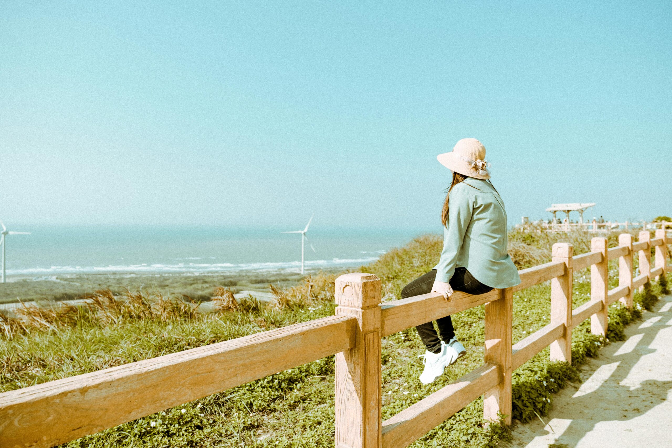 A woman in a sun hat sits on a wooden fence, enjoying a scenic coastal view with wind turbines.
